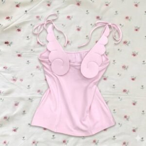 A light pink sleeveless camisole top with tie-up shoulder straps and decorative angel wing appliqués on the chest, displayed on a floral rose-print background