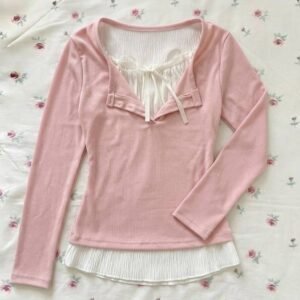 A pastel pink long-sleeved ribbed top featuring a white pleated fabric underlay at the neckline and hem, accented with a small white ribbon tie, displayed on a floral rose-print background