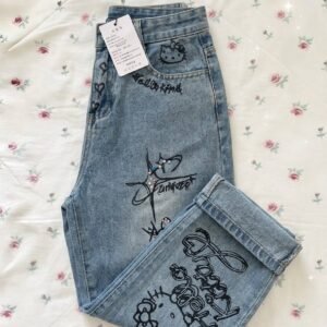 A pair of light-wash blue denim jeans featuring black Hello Kitty and doodle-style graphic prints on the pockets and legs, displayed folded on a floral rose-print background
