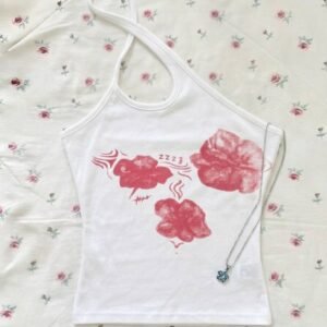 A white asymmetrical one-shoulder halter top with a red floral and abstract graphic print on the chest, shown with a blue flower pendant necklace on a rose-patterned background